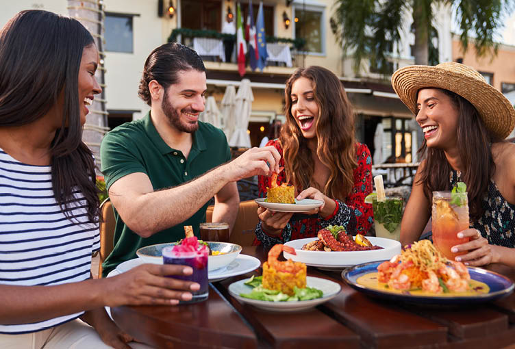 Vier Personen sitzen lachend an einem Restauranttisch im Freien mit Getr nken und Meeresfr chtegerichten.