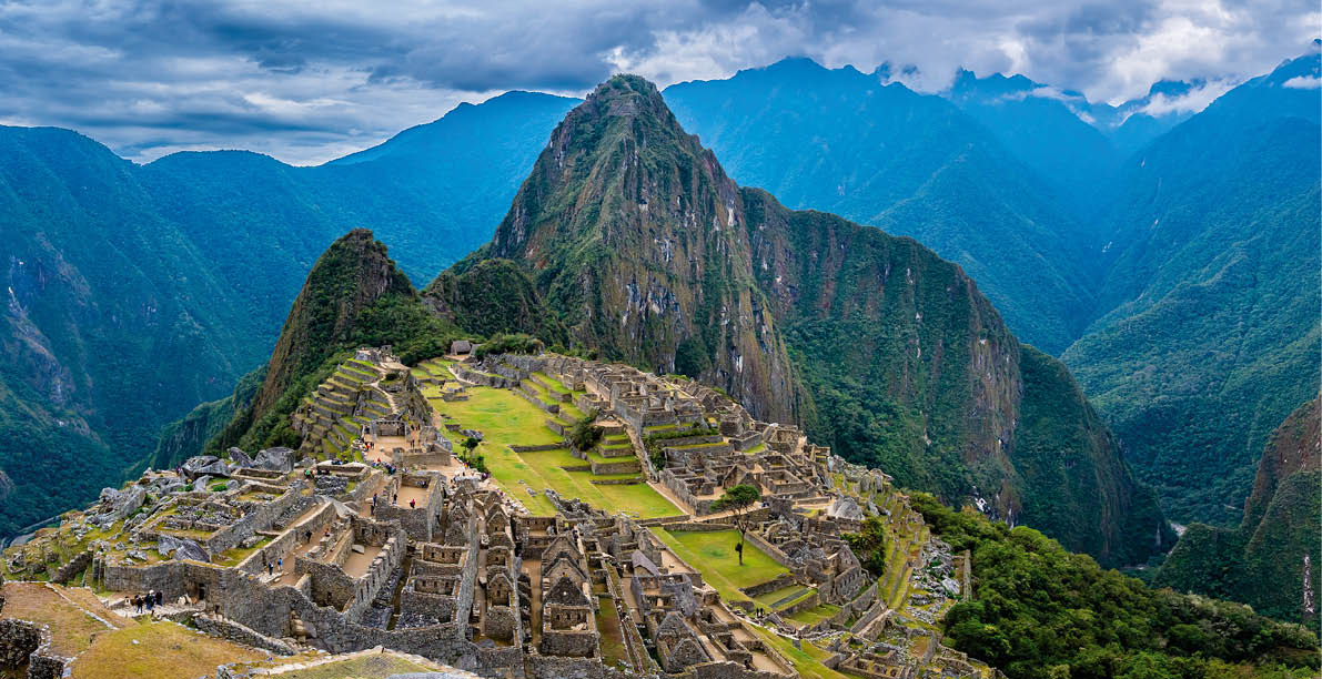 Panoramablick auf die Ruinen von Machu Picchu, umgeben von steilen, bewaldeten Bergen.