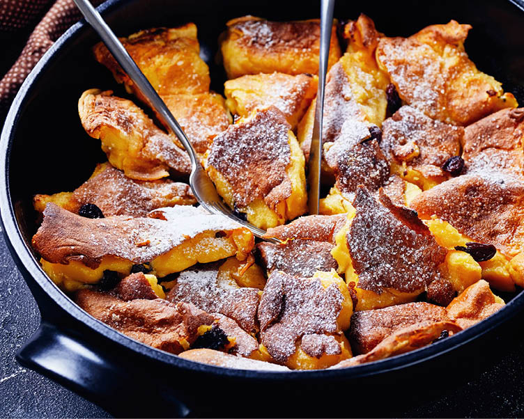 Light and fluffy austrian pancake kaiserschmarrn with raisins and icing sugar on top, cutlery on a black baking dish on a dark concrete table, horizontal view, close-up