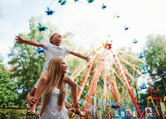 Daughter sits on the shoulders. Cheerful little girl her mother have a good time in the park together near attractions.