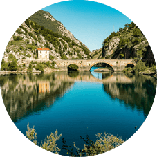 Alte Steinbr cke mit Rundb gen ber ruhigem See, umgeben von bewaldeten Bergen und zwei H usern.