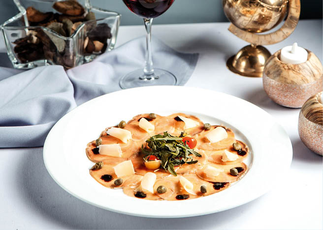 Feines Gericht mit Carpaccio, K se und Kapern, dazu ein Glas Rotwein, elegant angerichtet.