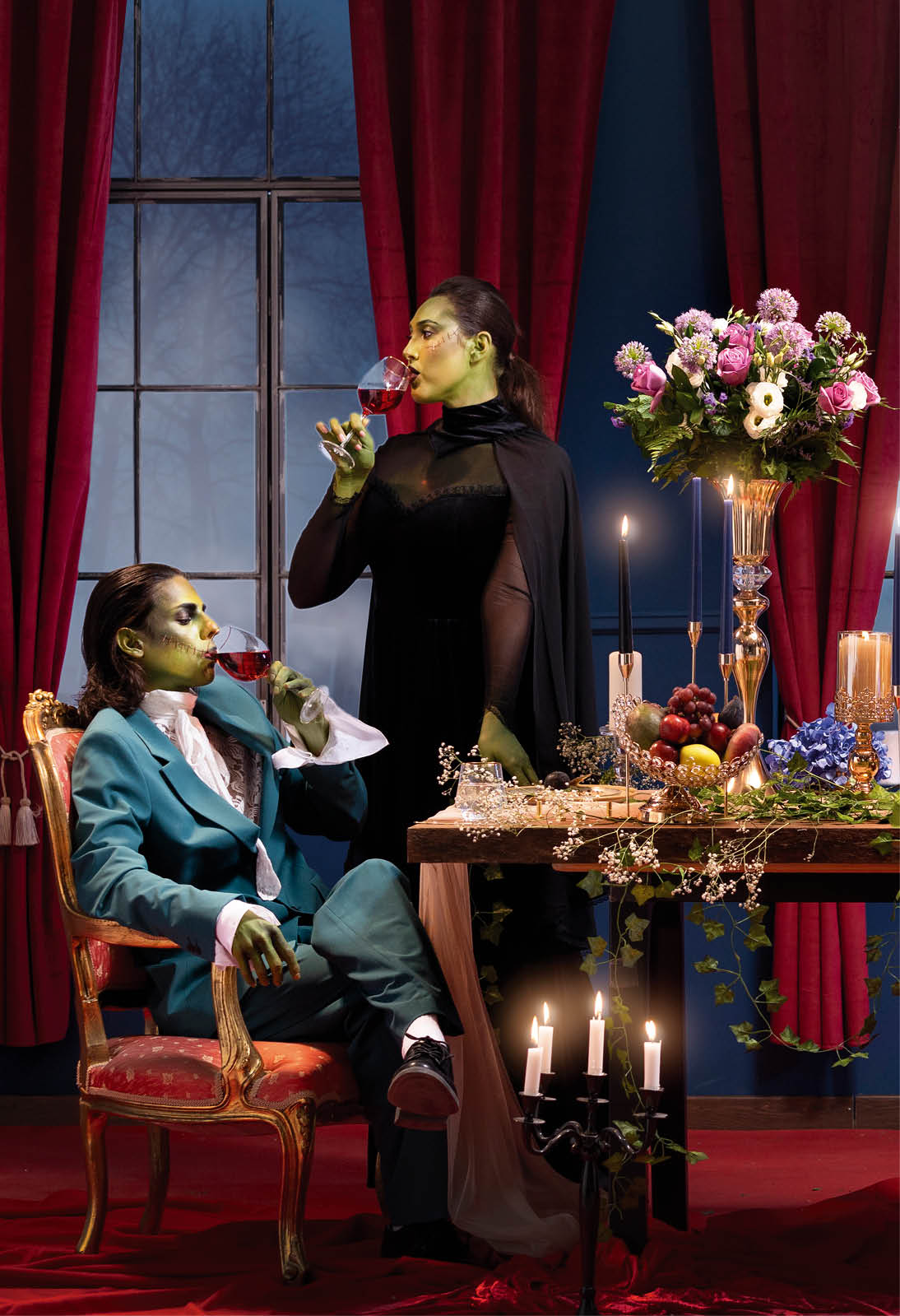 Zwei als Figuren mit gr ner Haut geschminkte Personen an festlich gedecktem Tisch mit Kerzen, Blumen und rotem Vorhang.