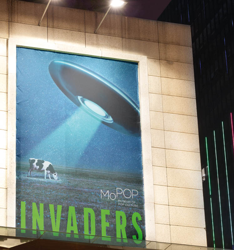 Leuchtplakat mit UFO ber grasender Kuh an Fassade bei Nacht.