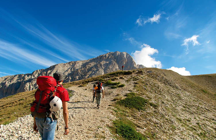 Gruppe von Wandernden mit Rucks cken auf steinigem Bergpfad vor felsigem Gipfel und blauem Himmel.