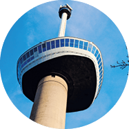 Aussichtsturm mit verglaster Plattform vor blauem Himmel, rechts ein kahler Baumzweig.
