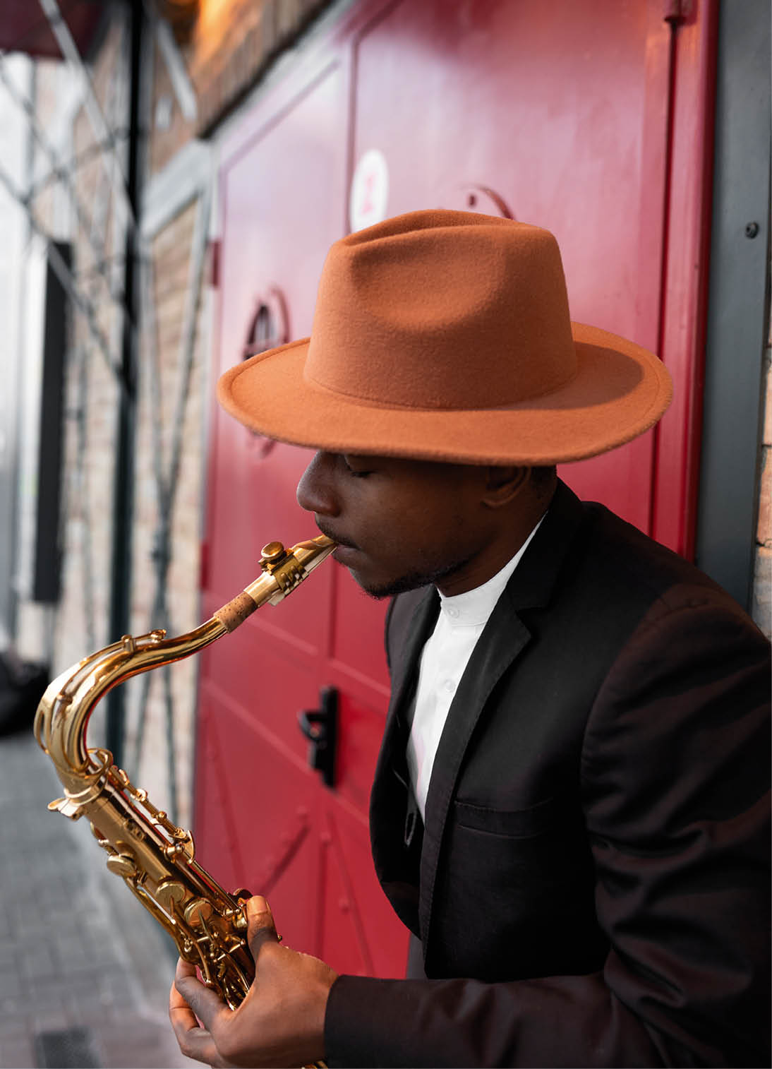 Mann mit braunem Hut spielt Saxofon vor roter T r.