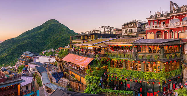Dicht bebaute H nge mit traditionellen H usern und Teeh usern, dahinter gr ne Berge und Meer bei Sonnenuntergang.