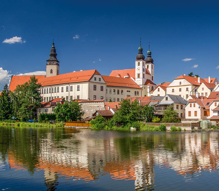 Rote D cher und Kircht rme spiegeln sich im ruhigen Wasser eines Flusses unter blauem Himmel.