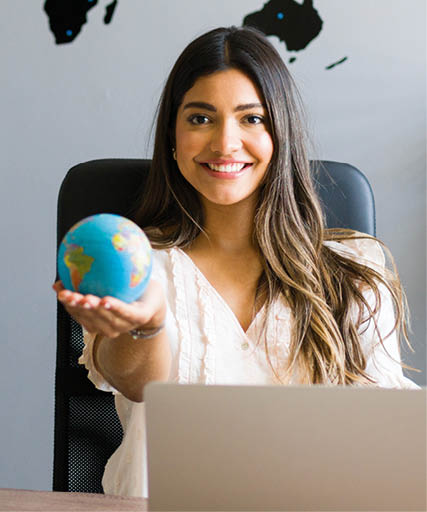 Das Bild zeigt eine junge Frau in einem modernen B ro, die mit einem L cheln einen kleinen Globus in der Hand h lt. Im Hintergrund sind eine Weltkarte an der Wand, eine Uhr, ein Globus auf einem Regal und verschiedene B romaterialien zu sehen – ein klassisches Reiseb ro- oder Bildungsumfeld. 