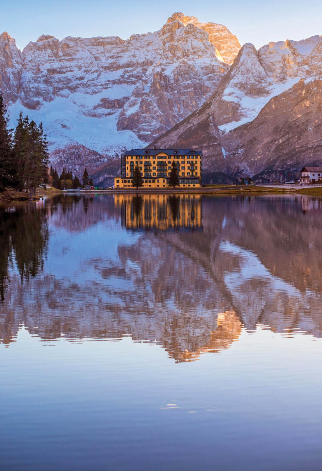 Hotel an einem See mit Spiegelung im Wasser, dahinter schneebedeckte Berge bei Sonnenaufgang.