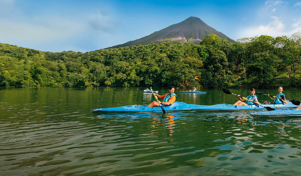Vier Personen paddeln in zwei Kajaks auf einem See mit bewaldetem Ufer und Vulkan im Hintergrund