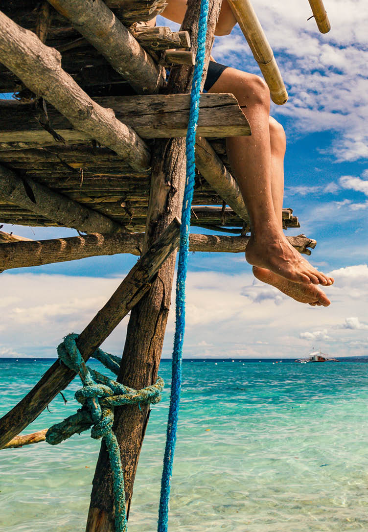 Person mit baumelnden Beinen auf Holzkonstruktion ber t rkisfarbenem Meer